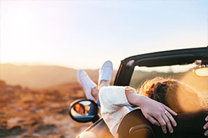 Eine Frau sitzt in einem Cabrio und hängt ihre Füße über die Autotür nach draußen. Mit ihren Händen hält sie sich am Sitz fest. Im Hintergrund sind verschwommen Berge und ein Sonnenuntergang zu erkennen.