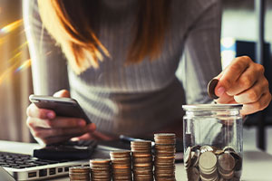 Eine Frau lehnt sich über einen Tisch. In der einen Hand hält sie ihr Handy und in der anderen Hand eine Geldmünze. Links am Bildrand ist ein aufgeklappter Laptop zu erkennen. Es sind fünf unterschiedlich große Stapel von Münzen zu erkennen. Rechts daneben ist ein Glas zu sehen, welches halb voll mit Münzen befüllt ist. 