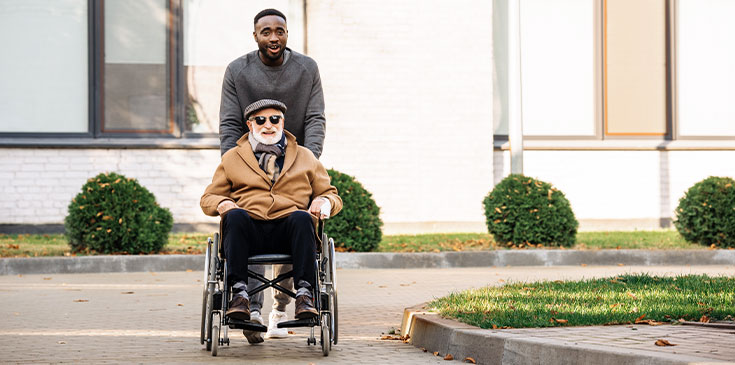 Neue Behinderten-Pauschbeträge ab 2021 Ein Mann in Pulli schiebt einen älteren Mann mit Jacke und Sonnenbrille in einem Rollstuhl über die Straße. Beide lächeln und sind gut gelaunt.