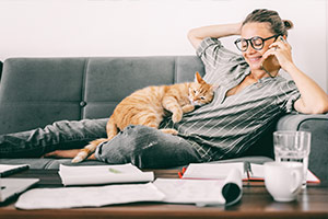 Eine Frau liegt auf einer Couch und ihre Katze auf ihr. Die Frau trägt eine Brille, sie telefoniert und lächelt. Auf dem Wohnzimmertisch vor ihr befinden sich ein Laptop, Unterlagen und Getränke.