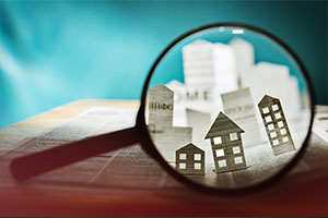 Vor einem blauen Hintergrund forciert eine Lupe eine Zeitung. Unter der Lupe werden Häuser, die aus der Zeitung „herausragen“ dargestellt.