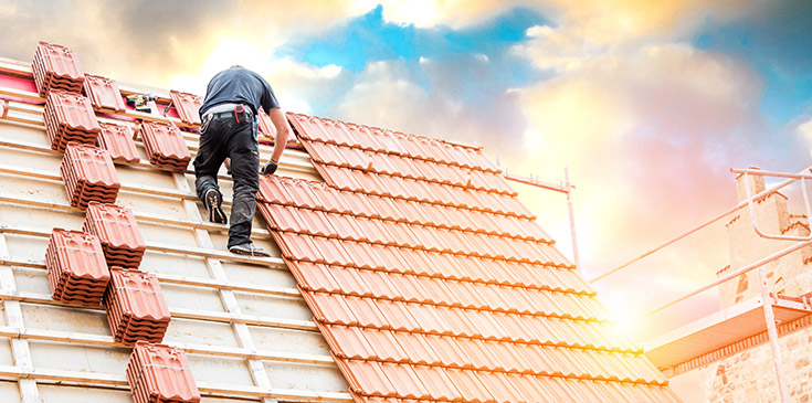 Ein Dachdecker arbeitete an einem Dach, das Dach ist teilweise nicht vollständig mit Ziegeln bedeckt. Im Hintergrund scheint die Sonne durch die Wolken.