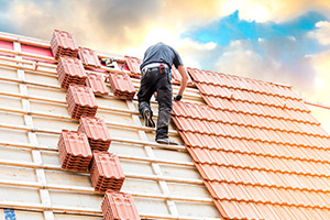 Ein Dachdecker arbeitete an einem Dach, das Dach ist teilweise nicht vollständig mit Ziegeln bedeckt. Im Hintergrund scheint die Sonne durch die Wolken.