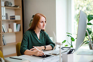 Eine junge Frau mit roten Haaren sitzt an ihrem Schreibtisch und blickt auf ihren Computer. Sie hält einen Stift über einem Tablet, das auf ihrem Schreibtisch liegt. Im Hintergrund befinden sich Pflanzen und Bücherregale.