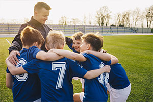 Ein Trainer und sein Team aus Jungs stehen im Kreis und haben die Arme über die Schultern der anderen gelegt. Alle lächeln.