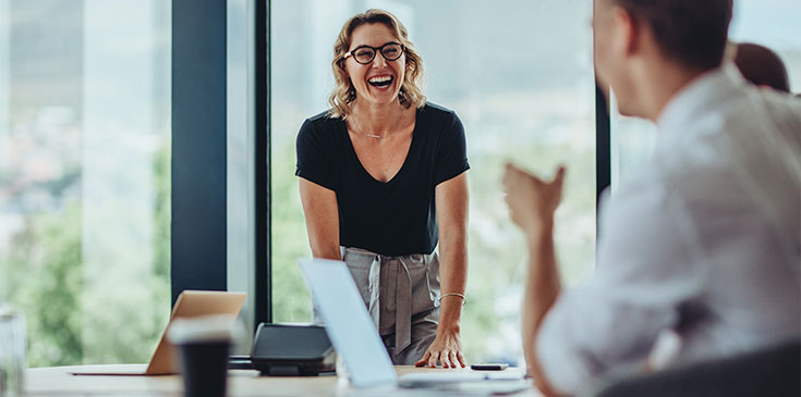 Eine Frau mit blonden Haaren und Brill steht im Büro an einem Tisch. Sie hat die Hände auf den Tisch gelehnt und lacht. Im Vordergrund des Bildes sind noch andere Personen verschwommen zu erkennen.