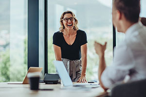 Eine Frau mit blonden Haaren und Brill steht im Büro an einem Tisch. Sie hat die Hände auf den Tisch gelehnt und lacht. Im Vordergrund des Bildes sind noch andere Personen verschwommen zu erkennen.