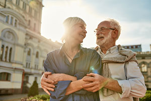 Eine ältere Frau und ein älterer Mann stehen vor einem historischen Gebäude. Sie lächeln sich an, haben die Arme umeinander gelegt und halten ihre Hände gegenseitig fest.