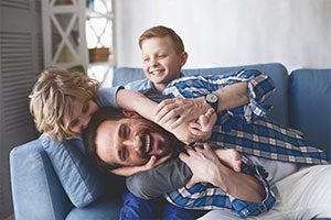 Ein Vater tobt mit seinen zwei Söhnen auf dem Sofa und lächelt in die Kamera.