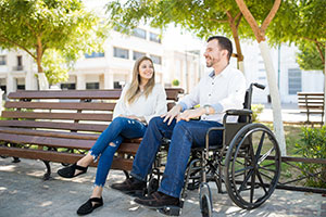 Ein Mann im Rollstuhl sitzt neben einer Frau, die auf einer Bank sitzt. Beide lächeln; im Hintergrund sind Bäume und Gebäude zu sehen.