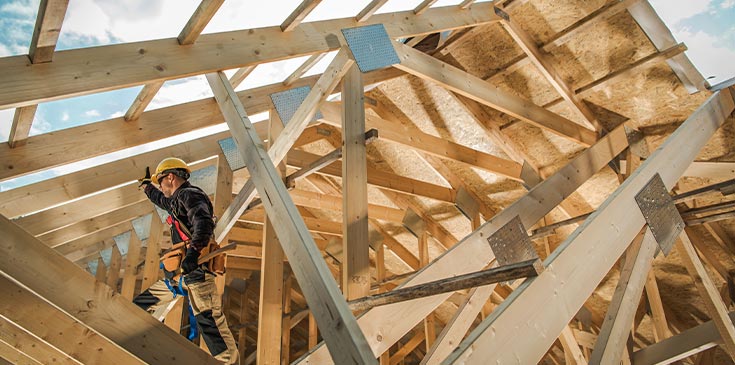 Sonderabschreibung: Steuervorteil für neue Mietwohnungen noch bis Ende 2021 sichern Ein Bauarbeiter mit Helm und Werkzeug steht auf einem Holzdach, das gerade ausgebaut wird. Durch das Dach hindurch sieht man den blauen Himmel