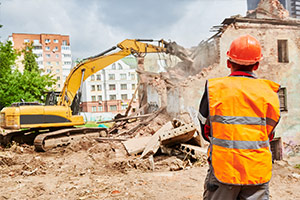 Ein Bauarbeiter steht mit dem Rücken zur Kamera und beobachtet, wie ein Bagger ein Haus abreißt.