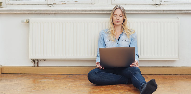 Energiepreispauschale 2022: Wer erhält sie und wann wird sie ausgezahlt? Eine junge Frau sitzt auf dem Boden vor der Heizung und schaut auf ihren Laptop.