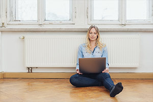Eine junge Frau sitzt auf dem Boden vor der Heizung und schaut auf ihren Laptop.