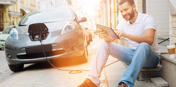 Elektromobilität: THG-Quote verkaufen und profitieren Ein junger Mann hält sitzend ein Tablet in der Hand während sein Elektrofahrzeug auflädt.