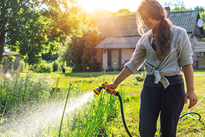 Eine junge Frau bewässert ihre Pflanzen mit einem Gartenschlauch, im Hintergrund steht ein Gartenhaus.