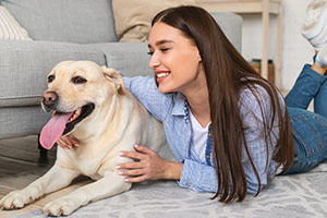 Eine junge Frau mit langen braunen Haaren liegt bäuchlings auf dem Fußboden. Neben ihr liegt ein cremefarbener Hund, ein Labrador – sie umarmt den Hund und lächelt ihn liebevoll an.