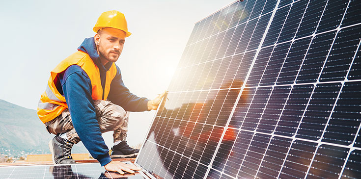 Photovoltaik und Steuern: PV-Anlagen bis 30 kWp steuerfrei, rückwirkend zum 1.1.2022 Ein Mann in Arbeitsmontur und mit gelbem Helm kniet vor zwei Solarpanels. Er ist dabei eine Photovoltaik (PV-Anlage) aufs Dach zu montieren.