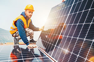 Ein Mann in Arbeitsmontur und mit gelbem Helm kniet vor zwei Solarpanels. Er ist dabei eine Photovoltaik (PV-Anlage) aufs Dach zu montieren.