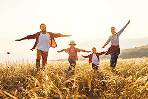 Eine Familie bestehend aus Mutter, Vater, Tochter und Sohn springt lachend über ein Feld während alle vier ihre Arme von ihrem Körper weg ausbreiten.