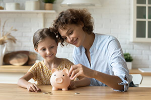 Eine Mutter und ihre kleine Tochter werfen lächelnd Geldmünzen in ein rosafarbenes Sparschwein.