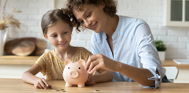 Eine Mutter und ihre kleine Tochter werfen lächelnd Geldmünzen in ein rosafarbenes Sparschwein.