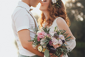 In der rechten Bildhälfte steht ein Hochzeitspaar, das sich anschaut und seine Gesichter aneinanderschmiegt. Die Braut trägt die Haare gewellt offen und hat Blumen im Haar, in der Hand hält einen Blumenstrauß. Der Bräutigam hat seine Hand auf die Hüfte der Braut gelegt.