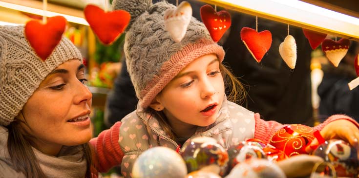 Eine Frau mit Wollmütze und Schal hat ihre kleine Tochter, die ebenfalls eine Wollmütze trägt, auf dem Arm. Beide sind auf dem Weihnachtsmarkt und schauen sich an einem Stand Weihnachtsschmuck an. Im Vordergrund hängen rote Herzen an einer Girlande. Das Kind staunt über die schönen Sachen und greift nach einer Christbaumkugel.