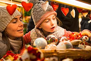 Eine Frau mit Wollmütze und Schal hat ihre kleine Tochter, die ebenfalls eine Wollmütze trägt, auf dem Arm. Beide sind auf dem Weihnachtsmarkt und schauen sich an einem Stand Weihnachtsschmuck an. Im Vordergrund hängen rote Herzen an einer Girlande. Das Kind staunt über die schönen Sachen und greift nach einer Christbaumkugel.