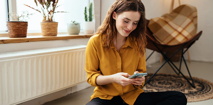 Energiepreispauschale für Studenten: Fristende für Antrag 2023 Eine junge Frau mit Smartphone in der Hand sitzt lächelnd vor der Heizung.