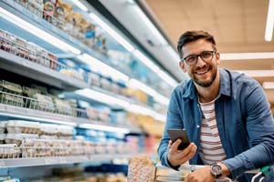 Eine Mann in Jeanshemd und geringeltem T-Shirt lehnt über einem Einkaufswagen. Der Einkaufswagen ist angeschnitten, man sieht nur den oberen Teil. Der Man hat ein Handy in der rechten Hand und schaut lächelnd in die Kamera. Aus dem Einkaufswagen ragt ein Weißbrot nach oben in den Sichtbereich. Im Hintergrund sind Einkaufsregale zu sehen. 