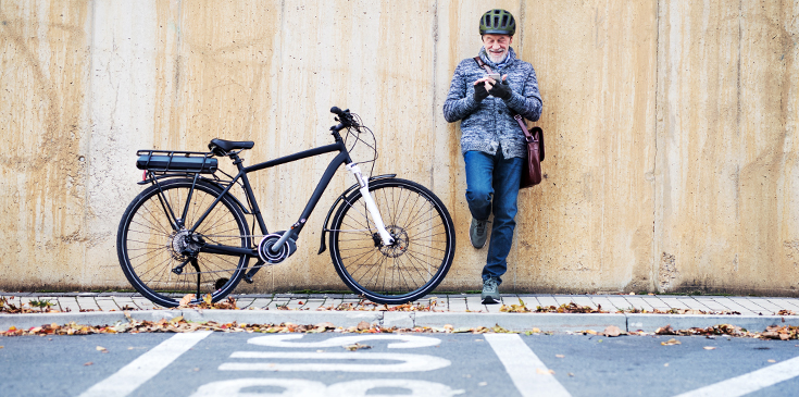 Ein älterer Mann steht auf einem Bürgersteig und lehnt sich mit dem Rücken an eine Wand, ein Bein ist angewinkelt. Er tippt etwas auf seinem Smartphone. Er trägt einen Fahrradhelm und eine Umhängetasche. Links neben ihm steht ein Elektro-Fahrrad. Auf der Straße vor ihm ist eine Spur mit der Aufschrift „BUS“ eingezeichnet.