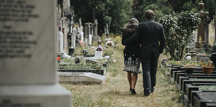 Eine Frau und ein Mann in schwarzer Kleidung laufen tagsüber über einen Friedhof zwischen Gräbern entlang. Sie sind von hinten zu sehen; der Mann hat seinen Arm um die Frau gelegt. Rechts und links von dem Paar befinden sich Gräber, Blumen und Gestecke.