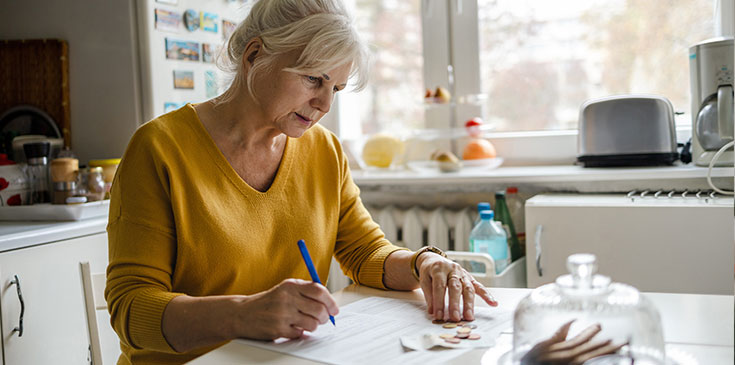 Freistellungsauftrag: Wie spare ich die Steuer auf Kapitalerträge? Eine Dame sitzt an ihrem Küchentisch, hat Dokumente sowie Bargeld vor sich und füllt ihren Freistellungsauftrag aus.