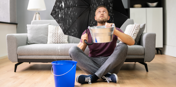 Hochwasserschäden steuerlich absetzen Mann sitzt im Wohnzimmer und fängt Wasser auf, das von der Decke kommt.