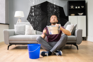 Mann sitzt im Wohnzimmer und fängt Wasser auf, das von der Decke kommt.