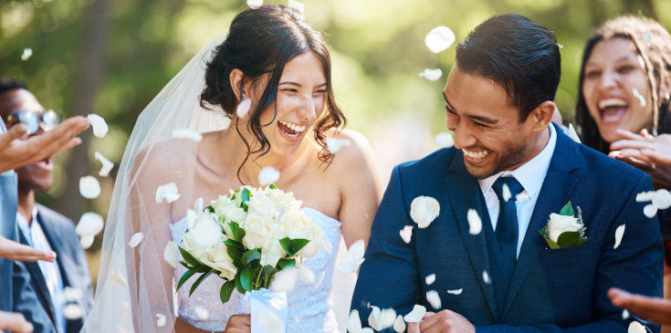 Nach der Hochzeit: Checkliste für Namenswechsel, Steuerklasse & Co. Ein frisch verheiratetes Paar lacht glücklich, während sie von Gästen mit Blumenblättern beworfen werden.