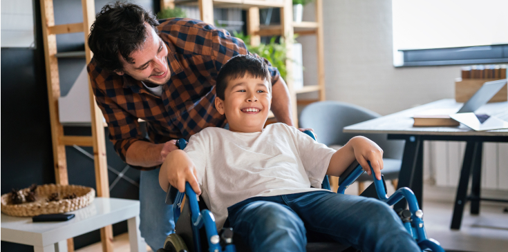 Steuervergünstigungen für Kinder mit Behinderung Ein Vater schiebt seinen Sohn mit einer Behinderung im Rollstuhl und macht Quatsch mit ihm.