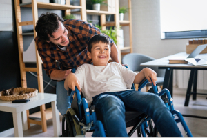 Ein Vater schiebt seinen Sohn mit einer Behinderung in einem Rollstuhl und macht Quatsch mit ihm.