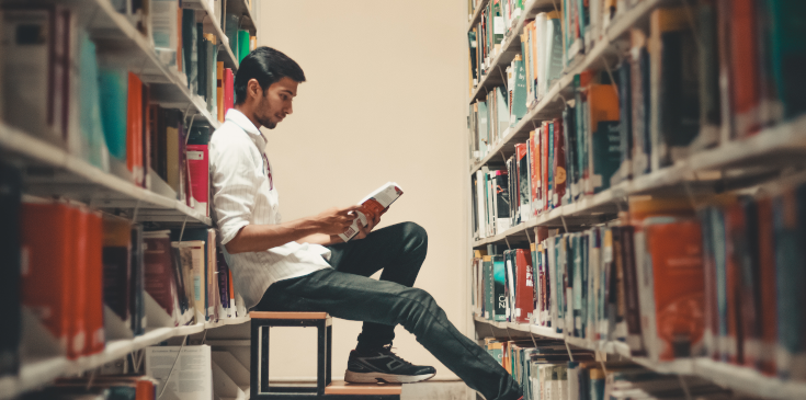 Wichtiger Unterschied in der Steuererklärung: Erststudium oder Zweitstudium? Ein junger Student ist in der Universitätsbibliothek und sitzt zwischen zwei großen Bücherreihen auf einem kleinen Tritt. Er trägt Jeans und Hemd und hält ein aufgeschlagenes Buch in der Hand, das er liest. Der Student ist von der Seite abgebildet, die Bücherreihen sind im Vordergrund verschwommen.