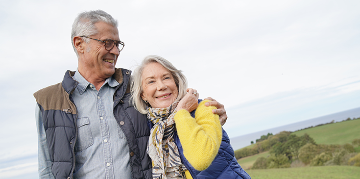 Ein älteres Paar steht im Freien, lächelt und umarmt sich liebevoll. Beide tragen warme Kleidung und wirken glücklich.