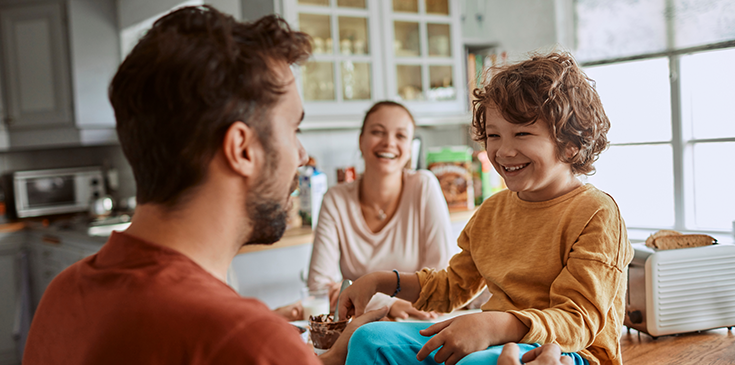Eine Familie, bestehend aus einer Frau, einem Mann und einem kleinen Jungen, sitzt in der Küche zusammen. Alle lachen. 