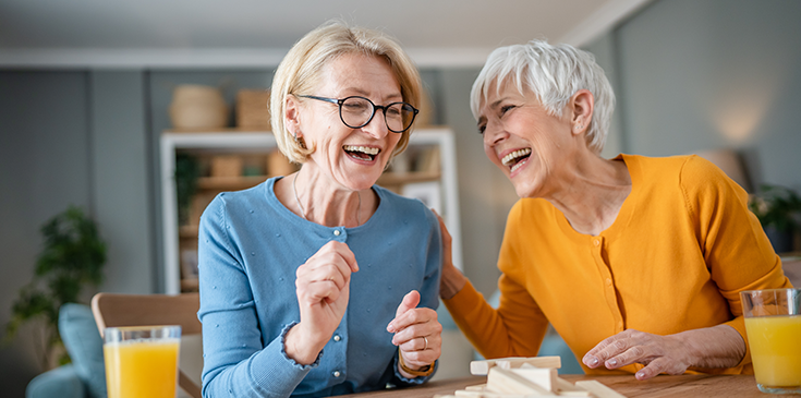 Zwei ältere Frauen lachen gemeinsam beim Spielen eines Gesellschaftsspiels an einem Tisch.