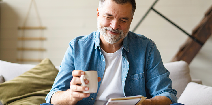 Lächelnder Mann mittleren Alters sitzt mit einer Tasse Kaffee auf dem Sofa und schaut auf sein Smartphone.
