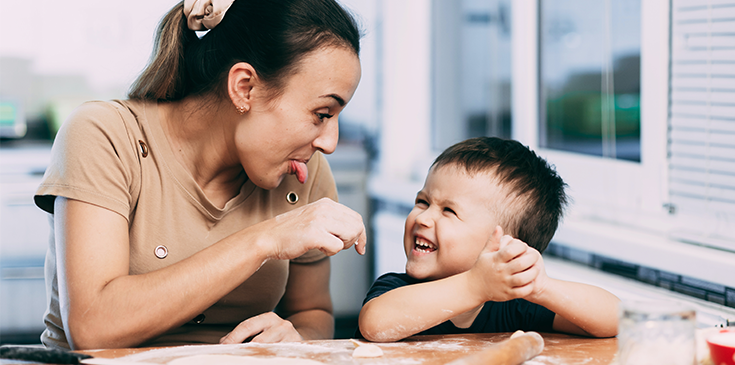 Eine junge Au-pair-Betreuerin sitzt gemeinsam mit einem lachenden Kind am Tisch. Beide lächeln sich an und verbringen eine fröhliche Zeit miteinander. Das Bild illustriert das Thema Au-pair-Aufenthalt und dessen steuerliche Grundlagen.