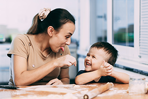 Eine junge Au-pair-Betreuerin sitzt gemeinsam mit einem lachenden Kind am Tisch. Beide lächeln sich an und verbringen eine fröhliche Zeit miteinander. Das Bild illustriert das Thema Au-pair-Aufenthalt und dessen steuerliche Grundlagen.