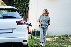 Eine Frau steht im Garten neben einem weißen Elektroauto, das gerade an einer Ladestation angeschlossen ist.