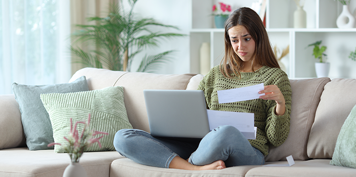 Junge Frau sitzt auf einem Sofa mit Laptop auf dem Schoß, hält ein Dokument in der Hand und schaut besorgt auf die Unterlagen.