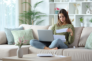 Junge Frau sitzt auf einem Sofa mit Laptop auf dem Schoß, hält ein Dokument in der Hand und schaut besorgt auf die Unterlagen.