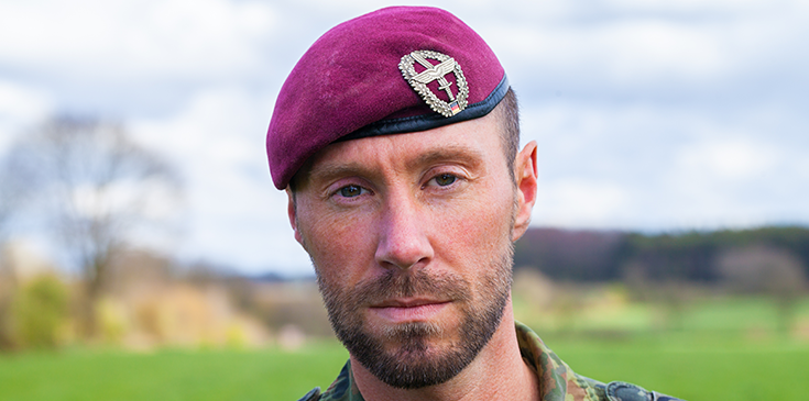 Soldat in Tarnuniform mit bordeauxrotem Barett blickt in die Kamera, im Hintergrund eine grüne Landschaft.